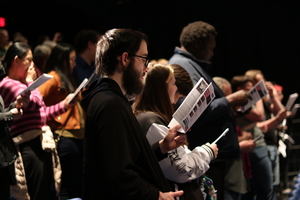 People standing and singing from sheets in a concert hall.