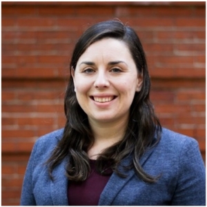 Smiling woman in a blue blazer against a brick wall.